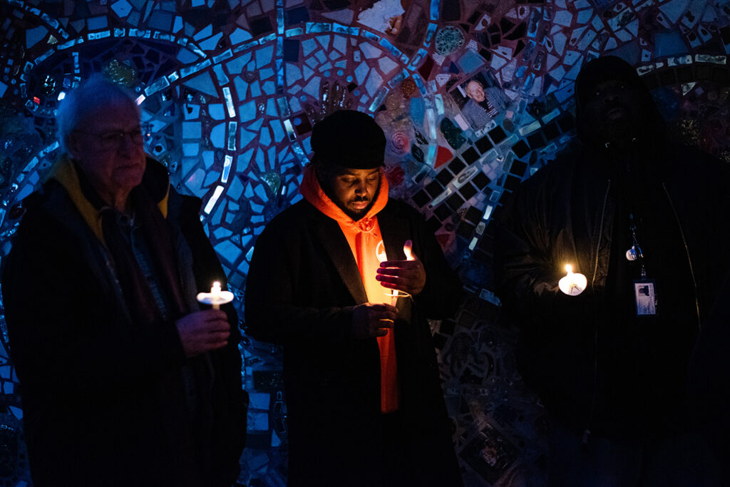 More people standing before a mosaic wall, holding lit candles. It is night time and the candles illuminate the people.