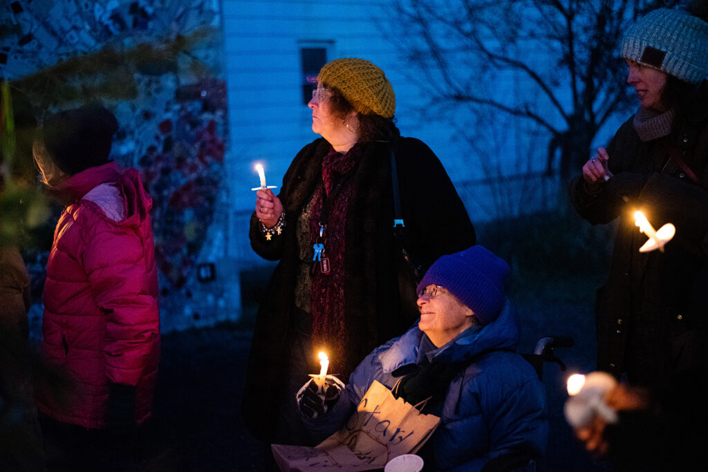 More gather around with lit candles in hand, featuring two people from a previous picture, still holding their signs and smiling as they prepare for the event.