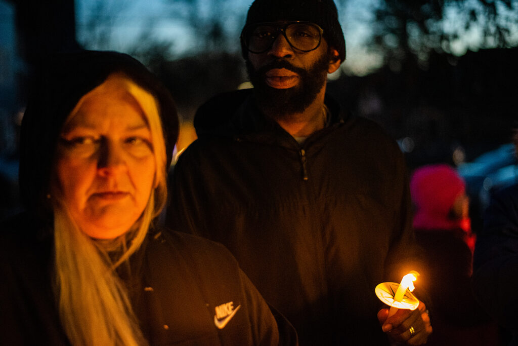 On the left, a light-skinned woman with long blond hair wearing a black hoodie. On the right, a person of color holding a li t candle. Both are looking at the camera.