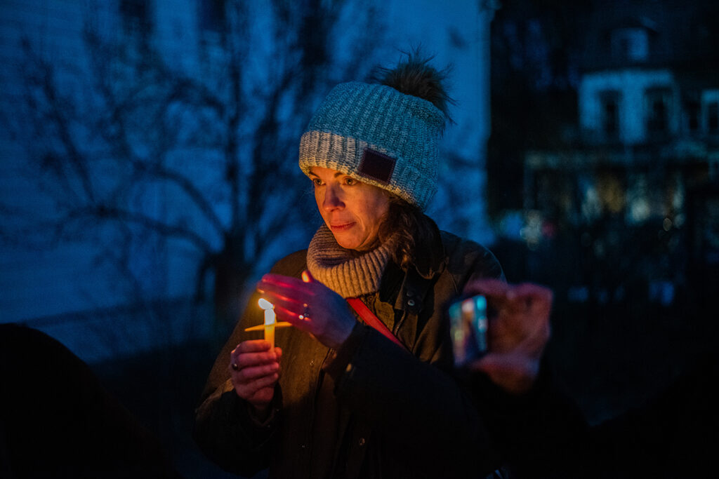 A light-skinned person wearing a grey beanie, holding a lit candle and having their hand over the flame to protect it. 