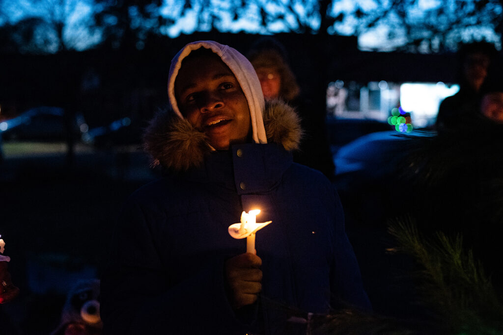 A single person of color holding a lit candle smiling at the camera, wearing a white hoodie and black puffer jacket.