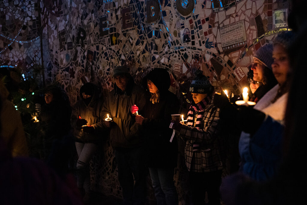 Sever people gathered against the mosaic wall, all holding and protecting their lit candles as they prepare for the event.