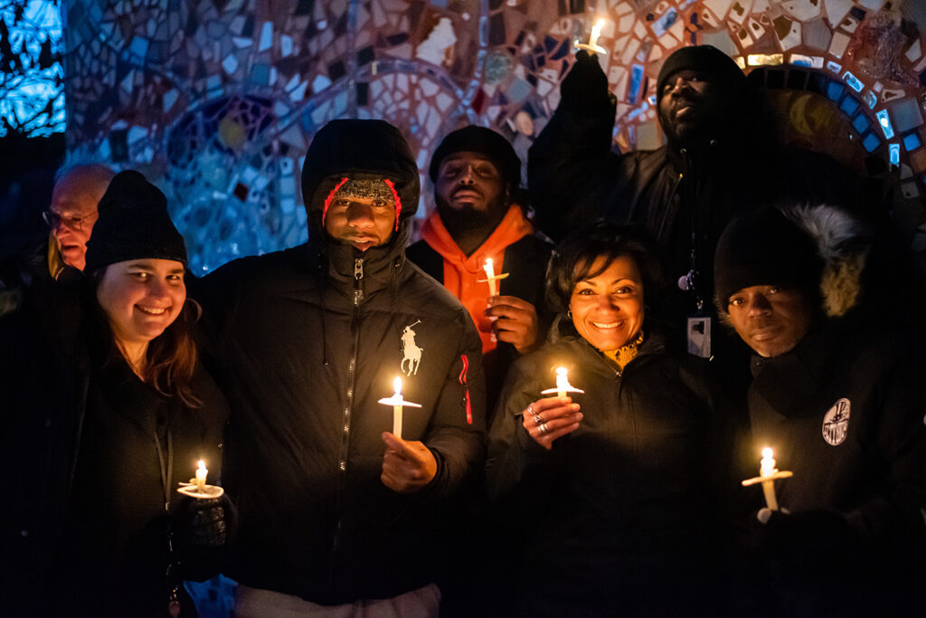 Several participants gather around with their lit candles, all smiling for the camera and holding their candles, standing In front of a mosaic wall. 
