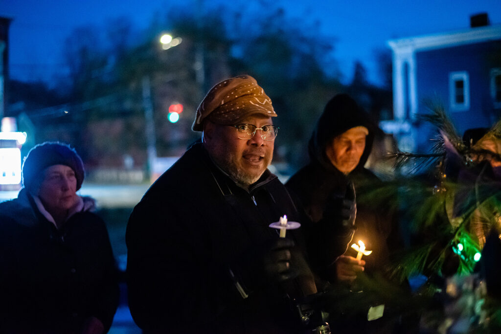 Three people standing before a tree. From left to right, a light -skinned person standing behind, and then two people of color holding their lit candles closer to the tree both wearing black hoodies.