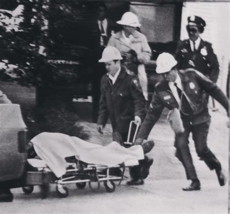 A black and white image with several officers and health officials gathered around a stretcher with a body covered in a white sheet. They seem frantic and are rushing to help.