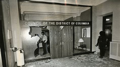 A black and white image with a large, somewhat destroyed wall with the words Council of the District of Columbia. Several officers are gathered around the chaos and examining the destruction, glass shattered on ground and hole in wall.