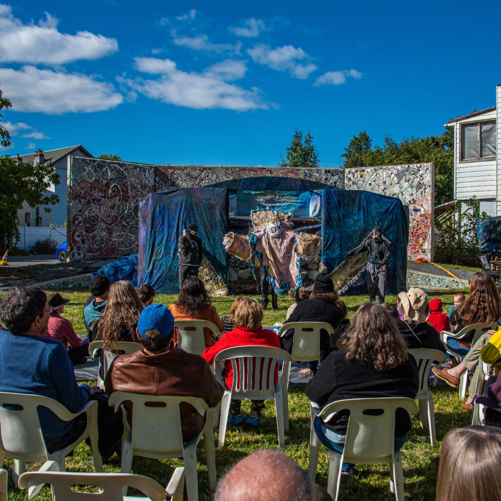 Blue skies over a the outdoor stage at Feedom Square, with a large puppet-based performance and a crowd watching from lawn chairs.