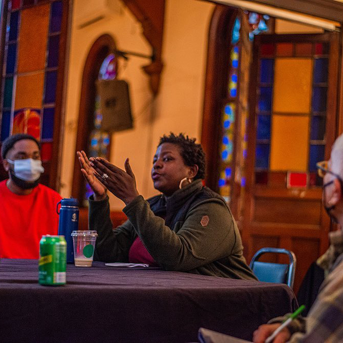 Yoruba Richen speaking to a group around a table at The Sanctuary for Independent Media, with stained glass windows in the background.