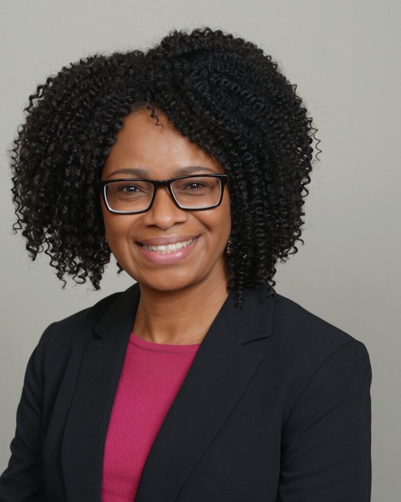 A head shot of Dr. Tina Omorogbe, a person of color wearing glasses with curly, short black hair wearing a pink shirt and a black blazer smiling for the camera.