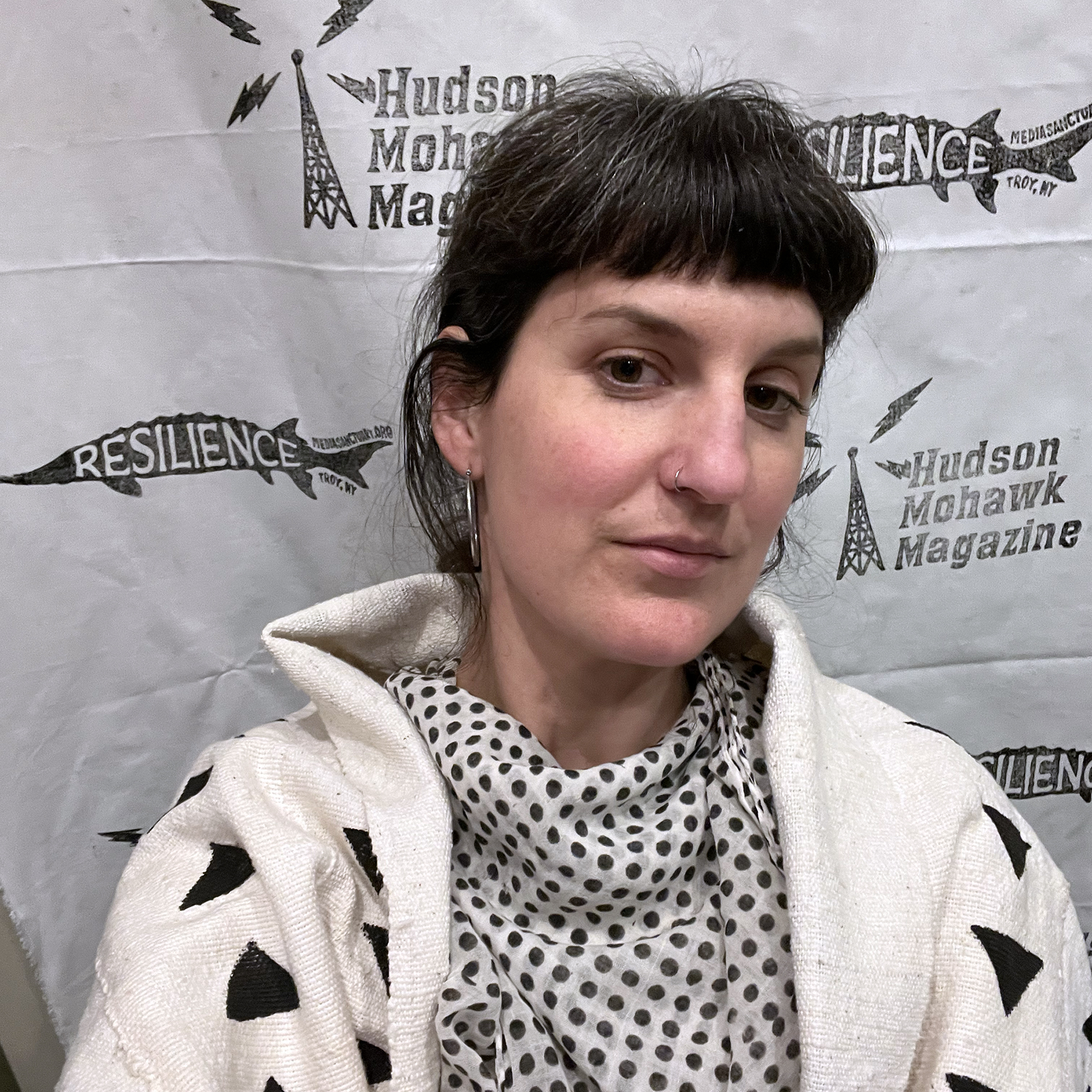 a portrait of Sina Basila Hickey in front of a black and white backdrop with the HMM and Sanctuary logo. Sina is a white woman with dark hair and bangs, a nose ring, hoop earrings, and is wearing black and white graphic patterned clothing.