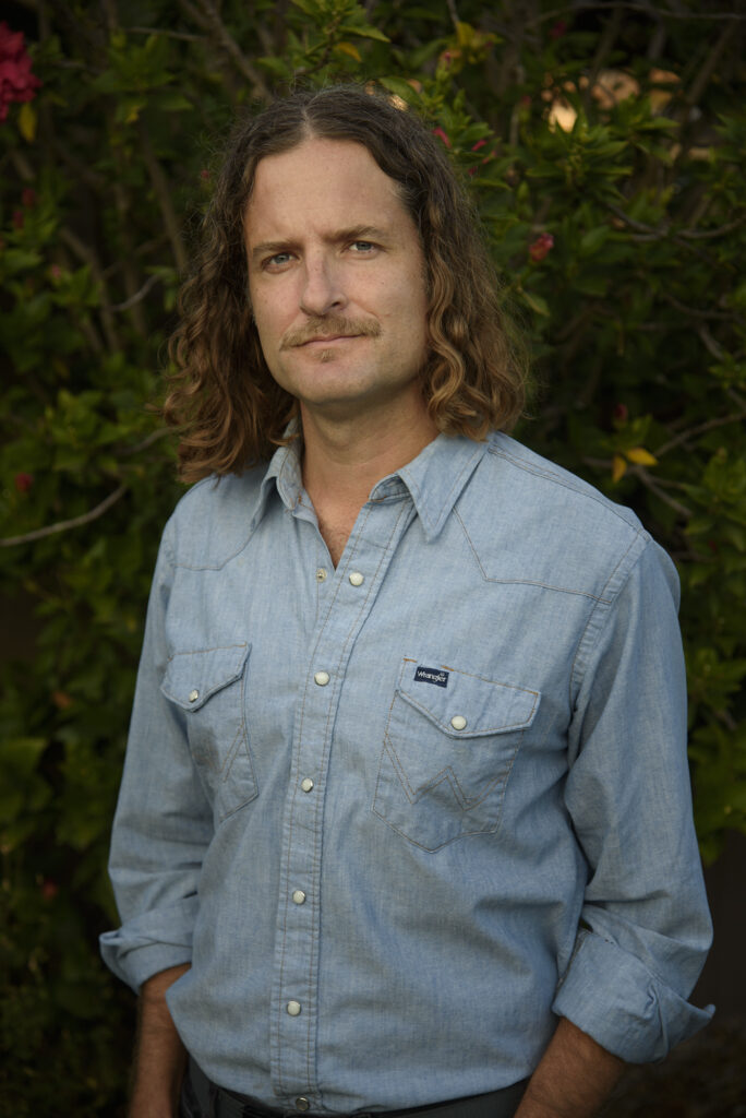 An upper-body headshot of Joshua Frank, who is wearing a blue button up and has medium-length brown, curly hair and facial hair. 