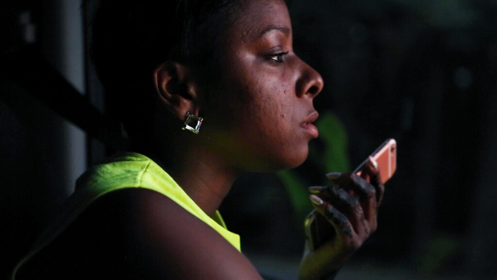 A photo of a person of color looking in the distance and holding a phone in front of a dark background, wearing a neon green shirt.