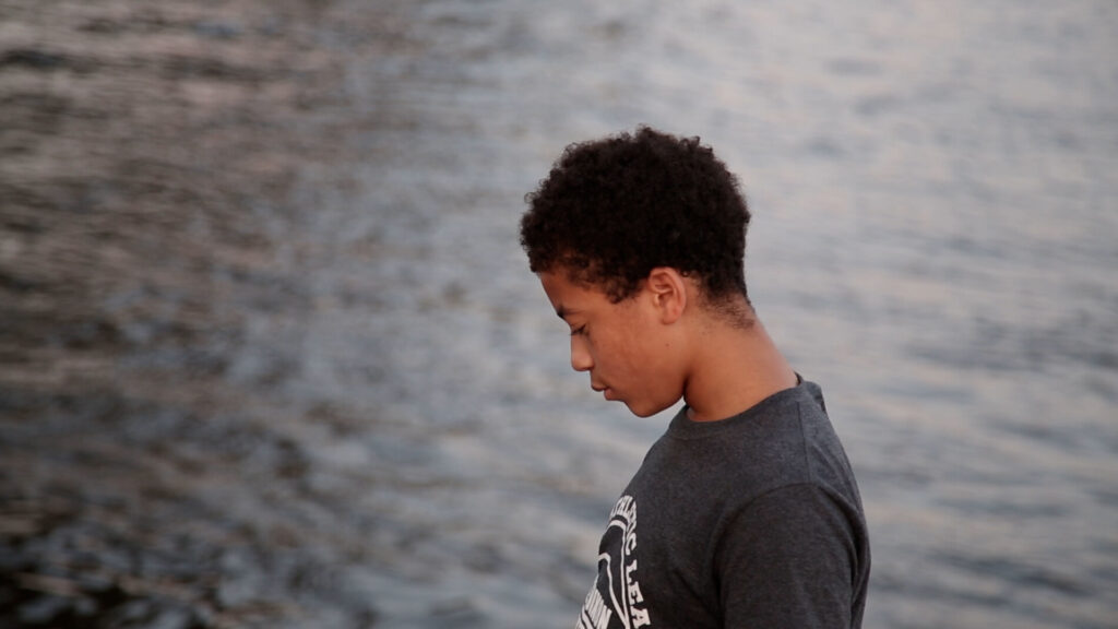 A photo of a young person of color standing in front of a body of water, looking down and off camera wearing a black shirt.