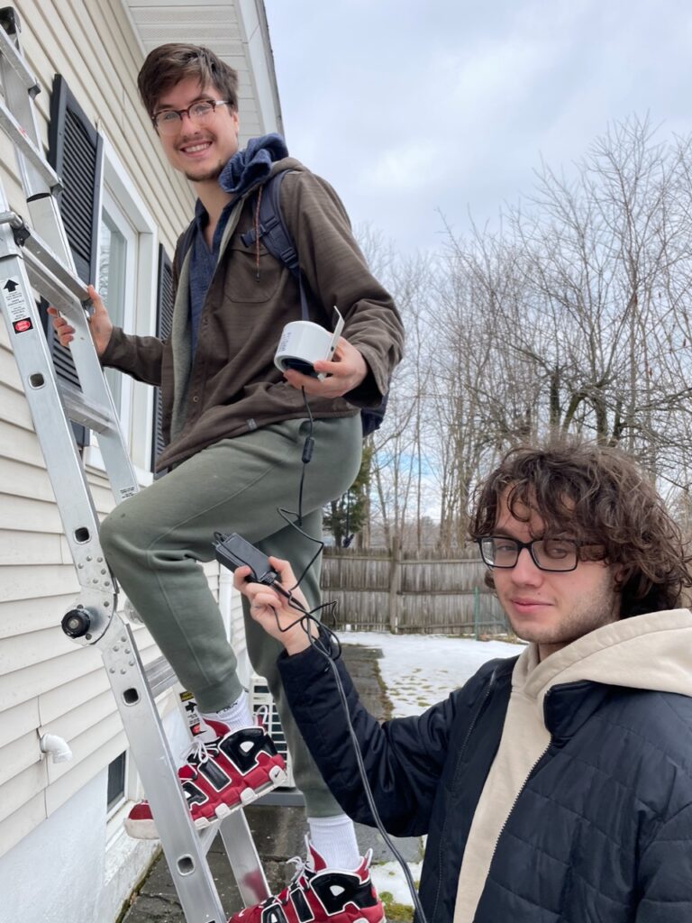 Two light-skinned young men install an air monitor on the side of a white house on a winter day. 