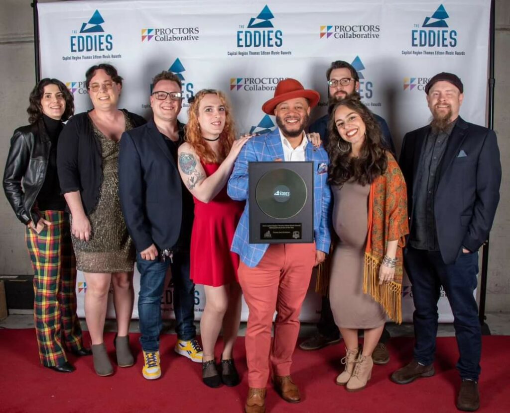 Members of the band Victory Soul Orchestra standing in front of a step and repeat banner on a red carpet with their Eddie Award