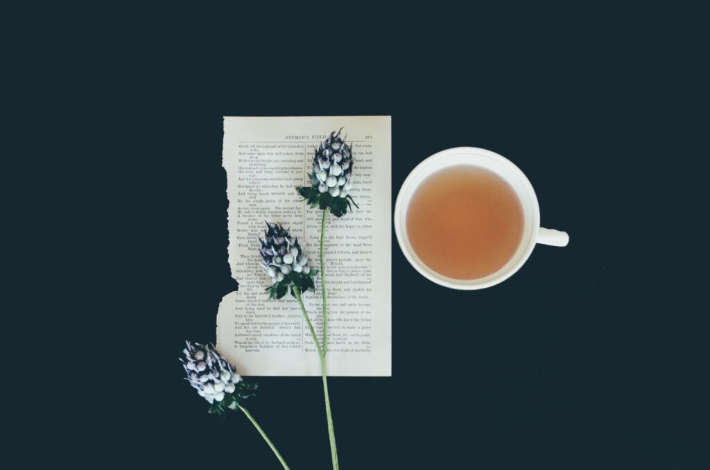 a cup of tea next to a strand of flowers on a page of a book. taken from above on a dark surfance