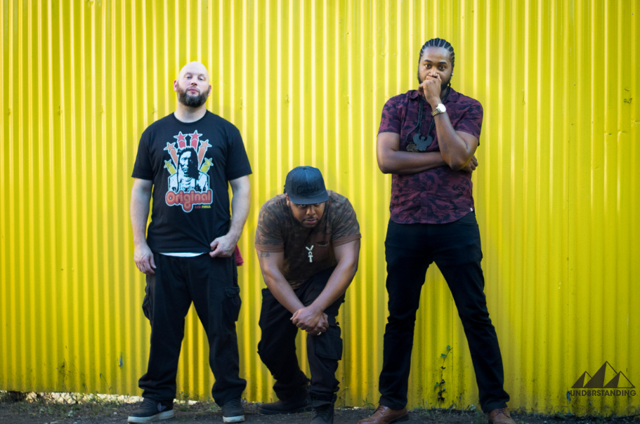 Three members of hip hop ground Und3rstanding pose in front of yellow wall. The person in the center is bent forward with hands on their knee