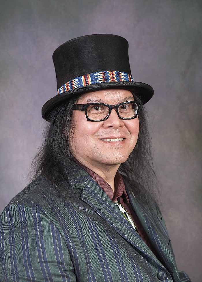 A headshot of Brent Michael Davids who is wearing a green-gray suit jacket, black framed glasses, and a top hat with an Indigenous band around it. He has dark hair to his shoulders.