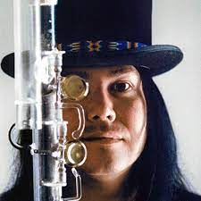 Composer Brent Michael Davids wears a top hat over long, straight, black hair in the background, with a glass flute in front of the left side of his face