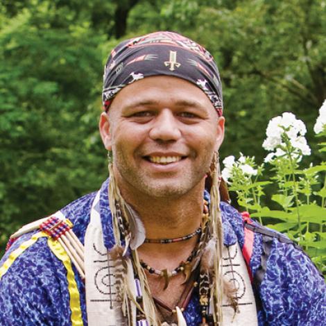 A medium close-up of Shawn Stevens (Red Eagle) of the Stockbridge-Munsee Band of Mohicans. He is wearing a purple shirt and has a headband on. He is smiling for the camera.