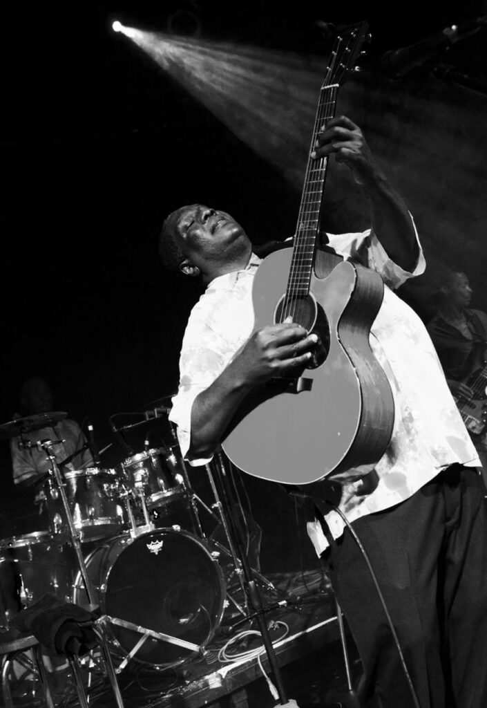 An action shot of Vusi Mahlasela, playing the guitar. The image is black and while, and there are a set of drums behind him while he palys.