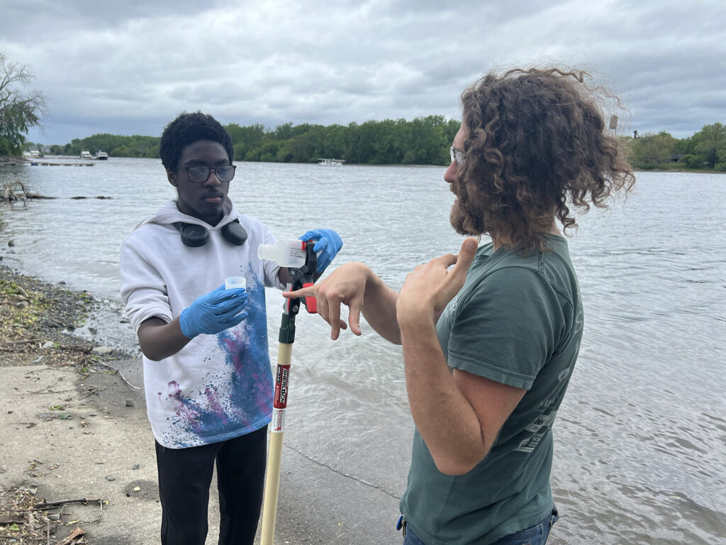 youth fellow Muzzamil wears blue gloves and holds a water sample at the end of sampling pole while mentor Sebastian looks on. Both stand on the shore of the Hudson River. 