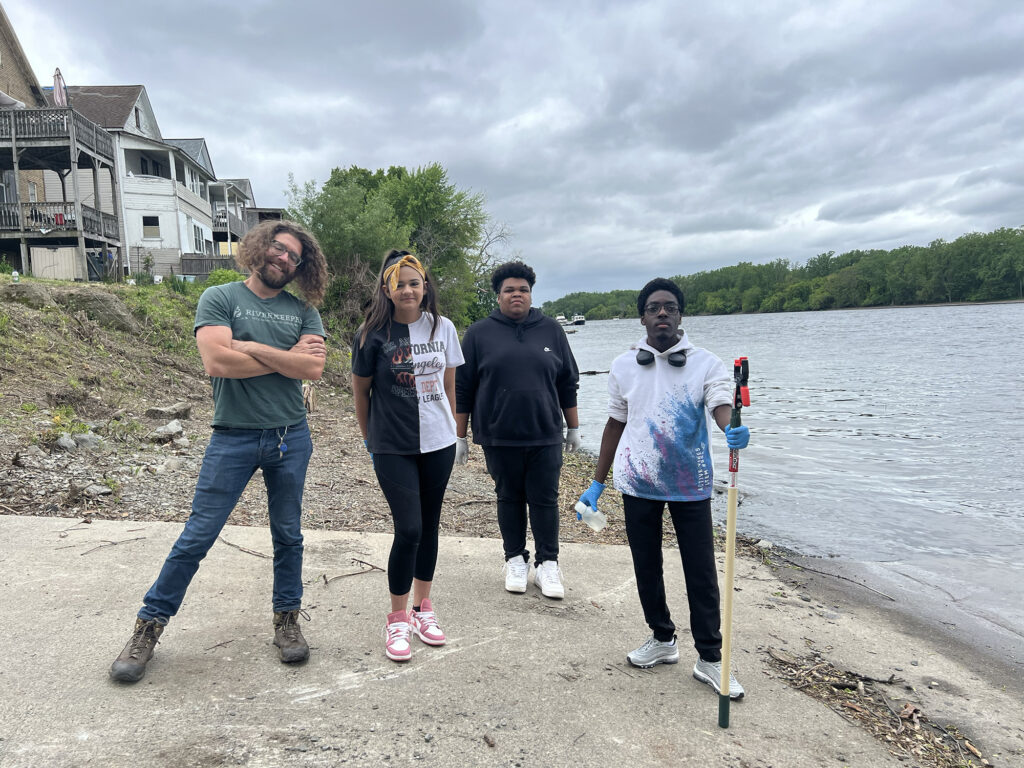 four people stand on the shore of the Hudson river on a concrete boat ramp, holding a testing pole and water sampling bottle. Three are youth fellows of high school age, one is a mentor.