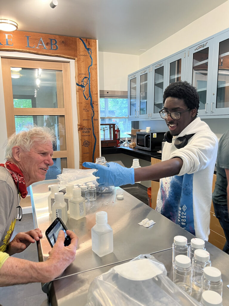 youth fellow Muzzamil working in the lab with volunteer Doug Reed. Muzzamil, wearing blue gloves, holds a vial of water while Doug examines it. A metal lab table with plastic bottles is between them.