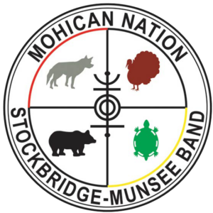 A circular symbol with the words "Mohican Nation, Stockbridge-Munsee Band" around the inside of the perimeter. There is a concentric circle within, which is divided into quadrants. Each quadrant contains the silhouette of an animal: a grey wolf on the top left, a brown turkey on the top right, a black bear on the bottom left, and a green turtle on the bottom right. 