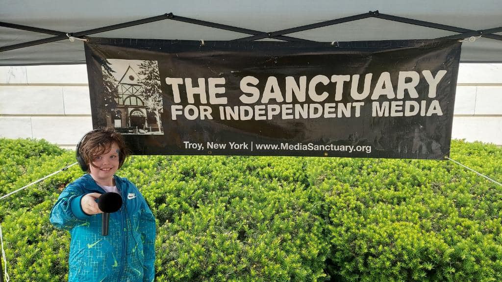 Matilda Rinker-Tennant holding a mic in front of the Sanctuary's banner.