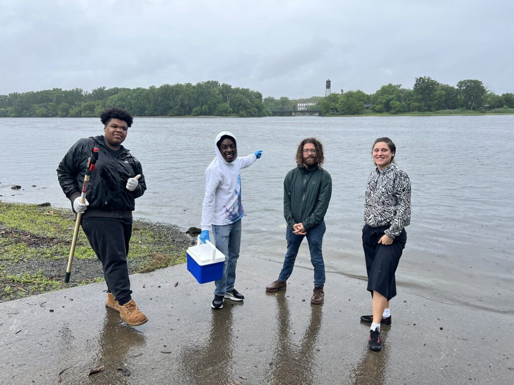 Two youth fellows and two mentors smile for a photo on the shore of the Hudson River on a rainy day. The youths are wearing vinyl gloves and carrying water sampling equipment. 
