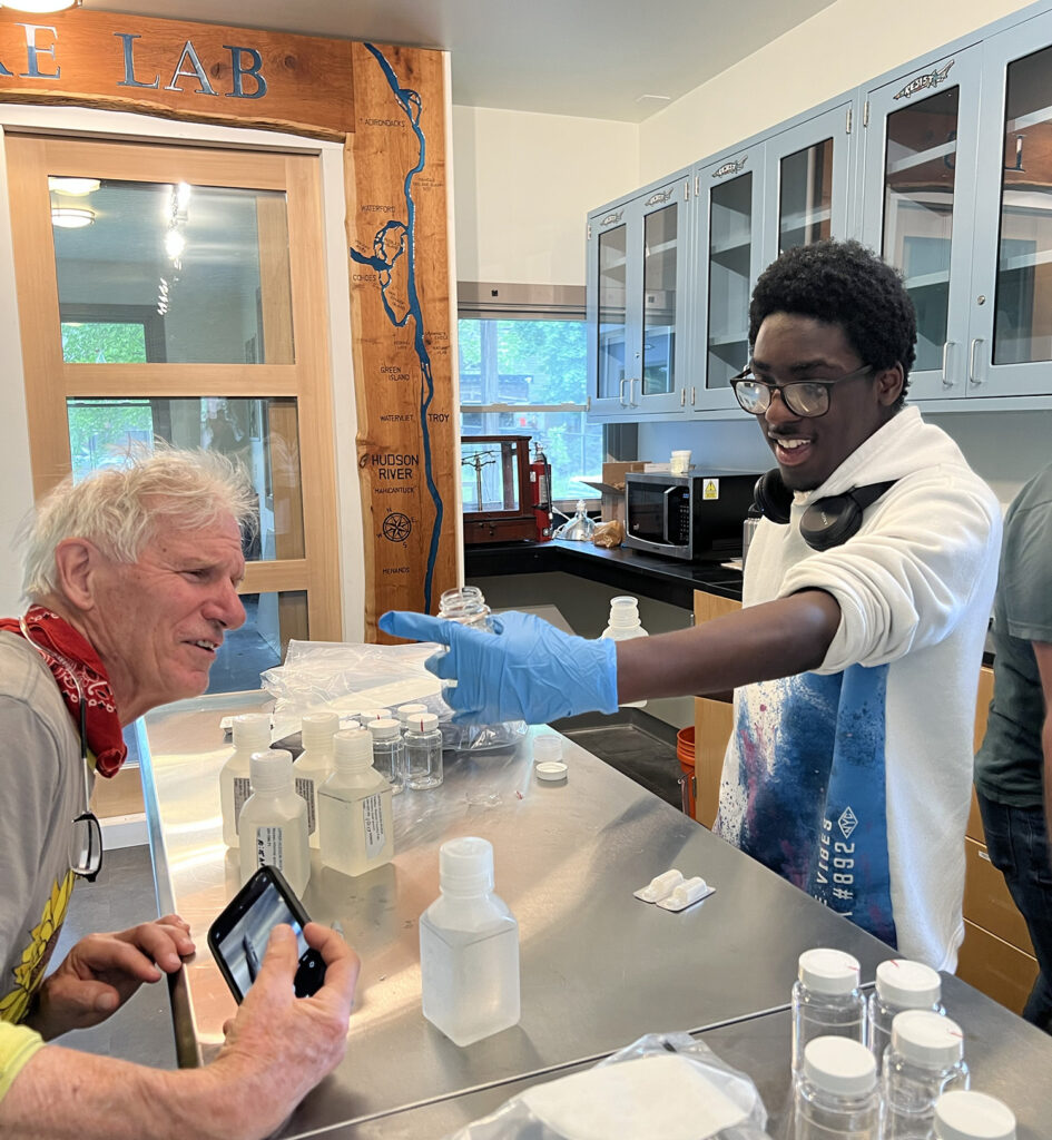 In NATURE Lab, a dark-skinned Sanctuary youth fellow wearing vinyl gloves proudly shows a water sample to  a light-skinned older man. 