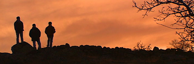 Three silhouetted figures on rocks looking at the sky which is orange and cloudy