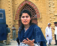 Sharmeen Obaid-Chinoy, with long dark hair and a dark blue scarf, looking into camera outside of Central Jail Karachi.