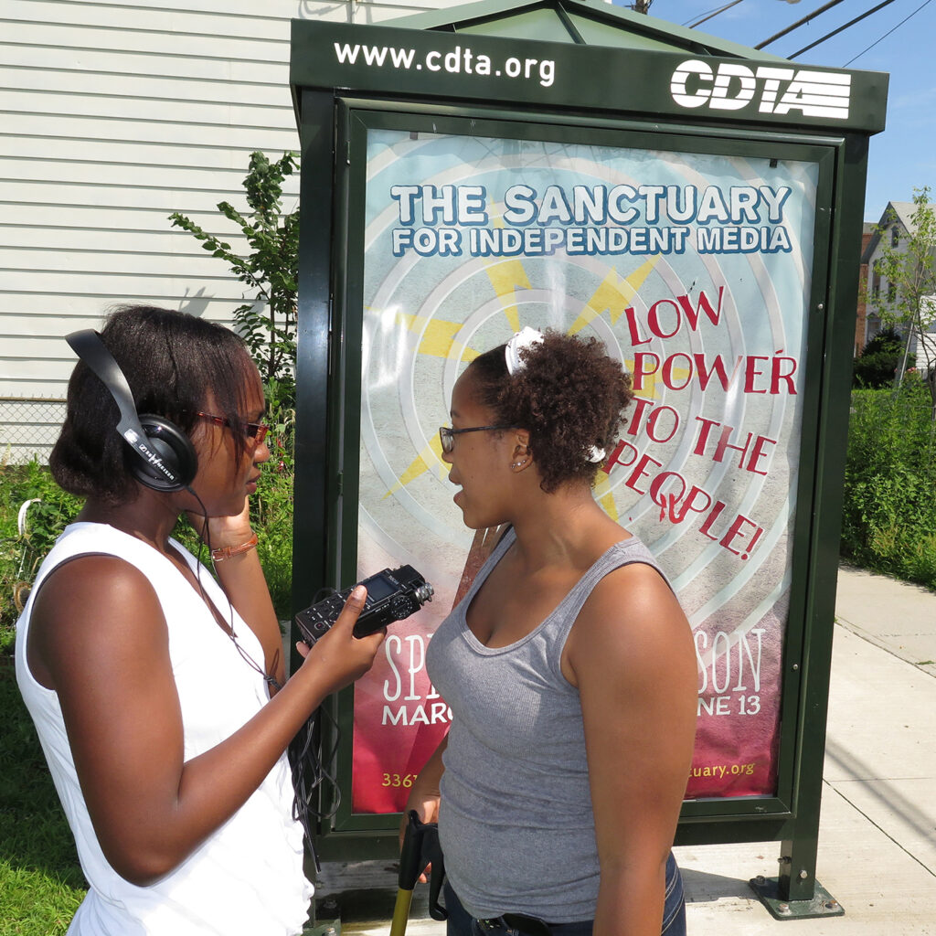 two young women do a radio interview in front of the bus stop