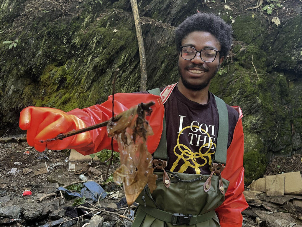 A Black teenage boy with glasses, standing in a low-flowing stream, smiles as he lifts a piece of film out of the stream with a stick. 