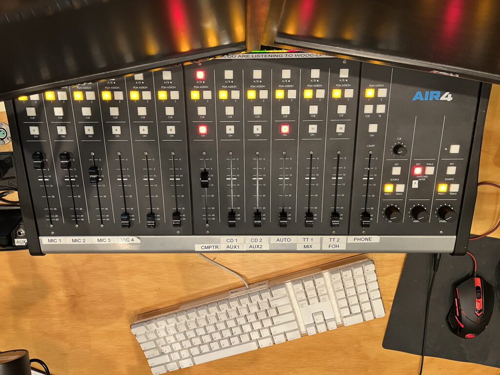 a photograph of the soundboard from above with the lights from the board reflecting on a black computer screen. In view is also a keyboard and mouse. 