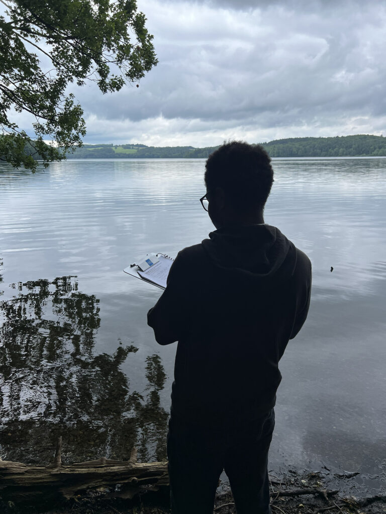 A young person, standing silhouetted against a body of water, takes notes on a clipboard. 
