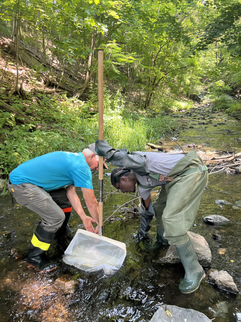 Two men wade in a creek and use tools to extract samples from the water. 