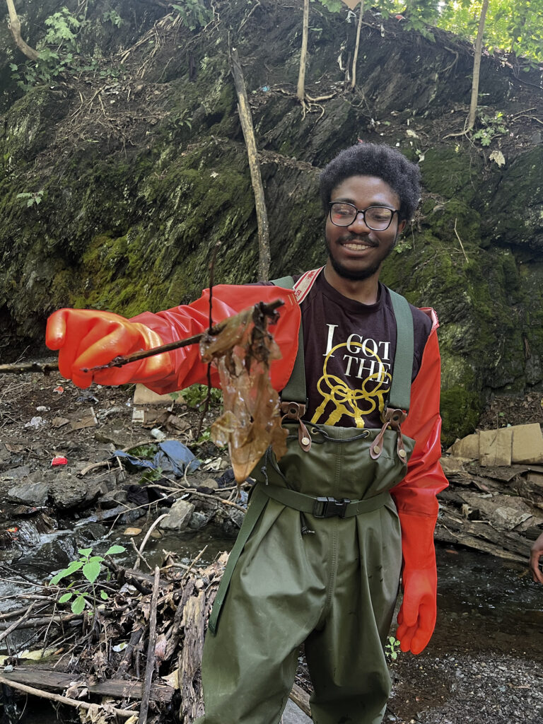 A Sanctuary youth ambassador, using a stick, lifts a thin brownish film out of a stream. He smiles at what he has found.  