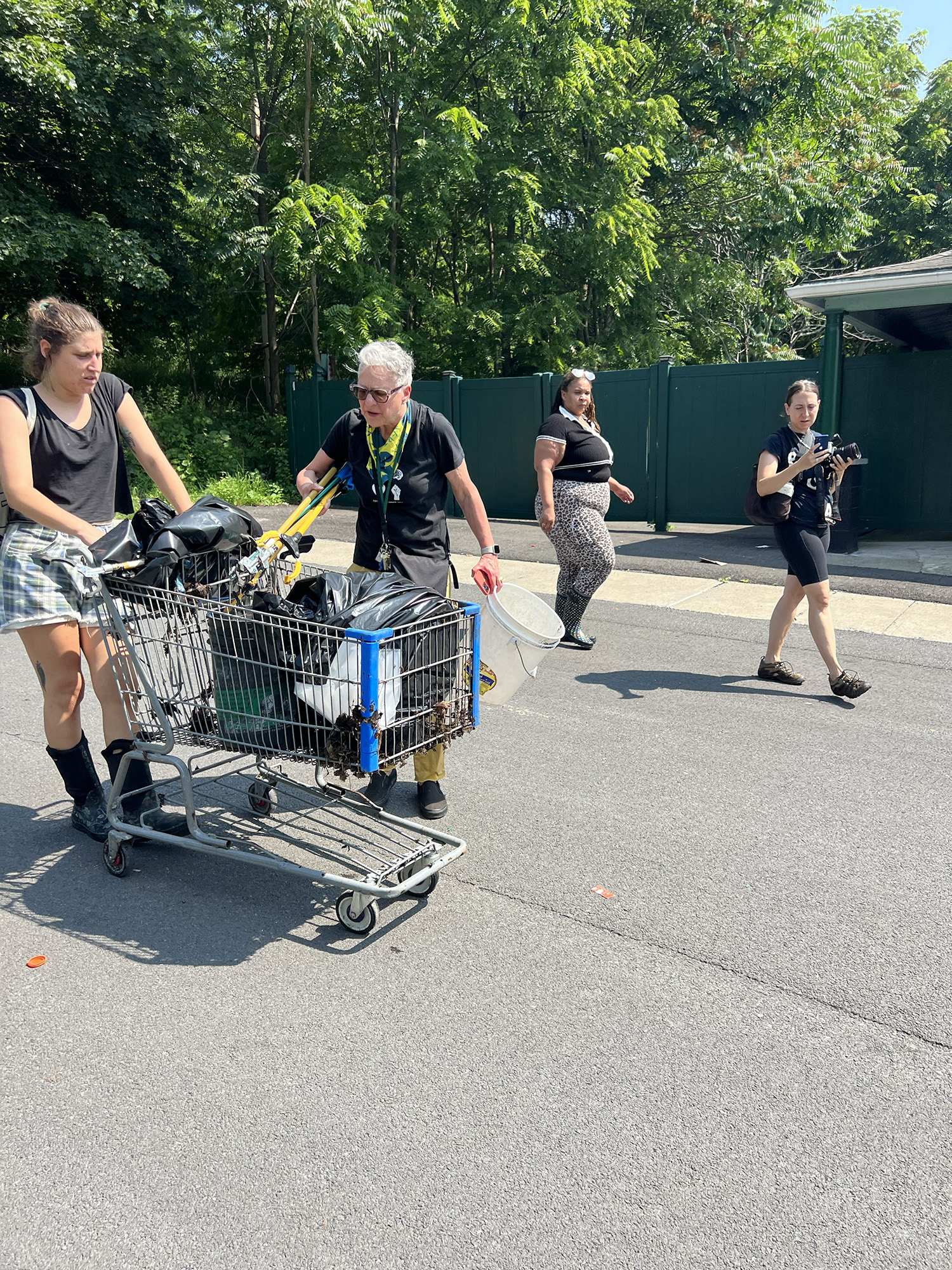 A woman pushes a shopping cart that contains what appears to be several bags of garbage. An older man carries a bucket and tools; two other women pass by in the background. 