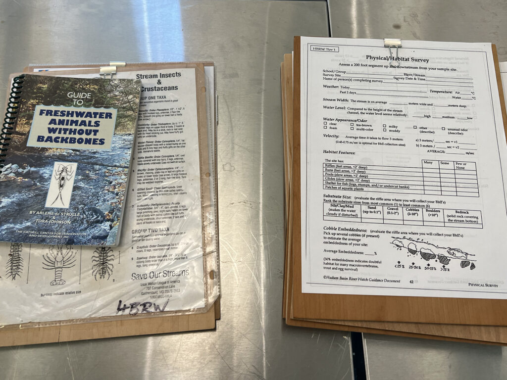 Two clipboards rest on a metal counter. The clipboard on the left contains information about freshwater invertebrates; the clipboard on the right holds a blank form for scientific notes. 