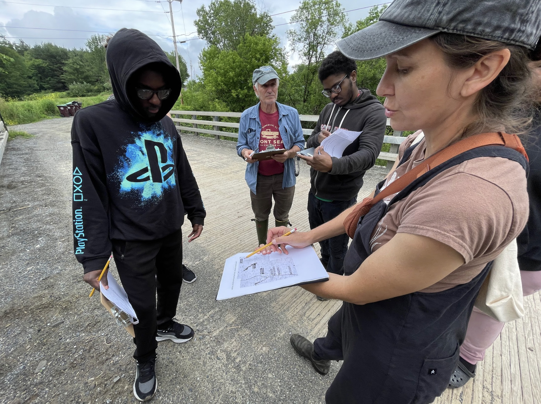 Ellie Irons, an older man, and two Sanctuary youth ambassadors stand on a bridge in a natural area, taking notes on clipboards. 
