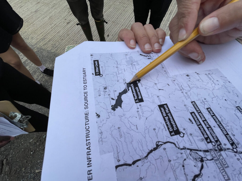 A close-up of the map of local water infrastructure. A light-skinned hand uses a pencil to point to the Tomhannock Reservoir on the map. 