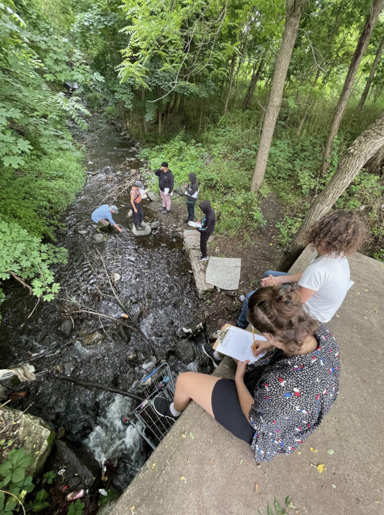Two youths sit on the edge of a culvert, taking notes, while five other people examine the low-flowing stream below. 