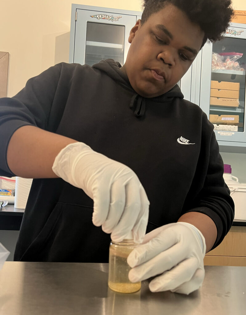 A Black teenage boy, wearing white vinyl gloves, examines a water sample in the NATURE Lab.