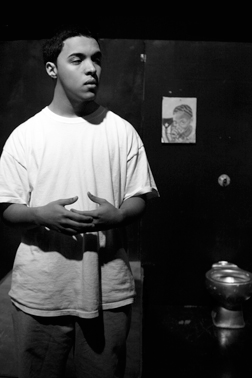 Image of a prisoner in a white tee and grey sweats. Posed with arms in front of his body, as if he is speaking. His facial expression is serious.