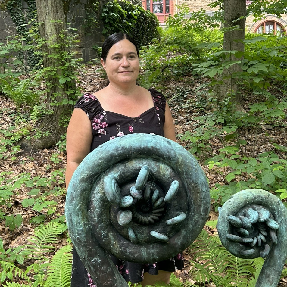 Misty Cook stands behind two large sculptures of curled fiddleheads of ferns shaped into spirals. the sculptures appear to be made of copper with a blueish-green patina on the top and dark gray underneath. the setting around Misty and sculptures in verdant green with trees overhead. Misty wears a brown sleeveless dress and has her brown hair pulled back.