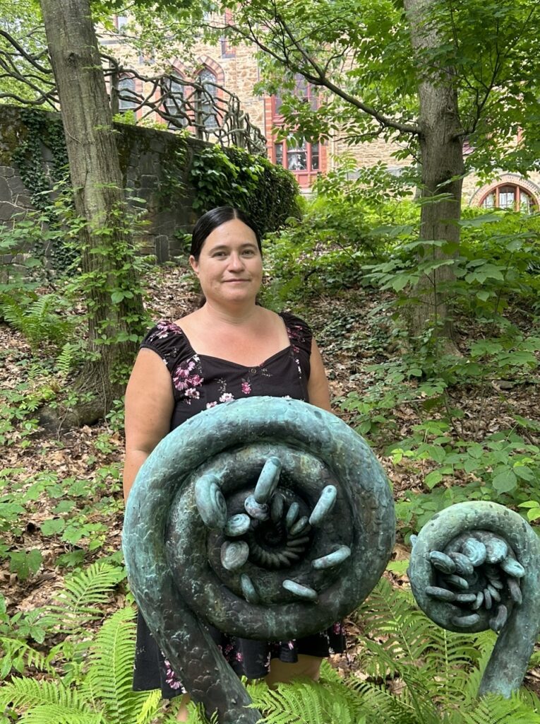Misty Cook stands behind two large sculptures of curled fiddleheads of ferns shaped into spirals. the sculptures appear to be made of copper with a blueish-green patina on the top and dark gray underneath. the setting around Misty and sculptures in verdant green with trees overhead. Misty wears a brown sleeveless dress and has her brown hair pulled back.
