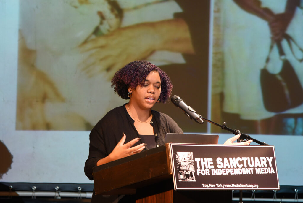 Jayana LaFountaine, an Afro-Latina photographer and doula, stands behind a podium at The Sanctuary for Independent Media with a photo slideshow depicting photos of her family projected behind